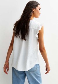 Vit ärmlös blus med en subtil texturerad finish, rynkdetaljer vid axlarna och en lös passform, kombinerad med ljusblå jeans.