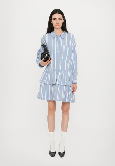 Helles blau gestreiftes Hemdkleid mit einem gestuften Volantsaum, ausgestattet mit einem Kragen und einer Brusttasche. Das Model hält ein schwarzes Accessoire.