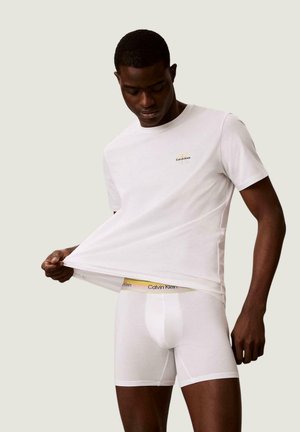 Uomo di colore con t-shirt bianca Calvin Klein e boxer bianchi, solleva la maglietta per mostrare la cintura con il nome del marchio.