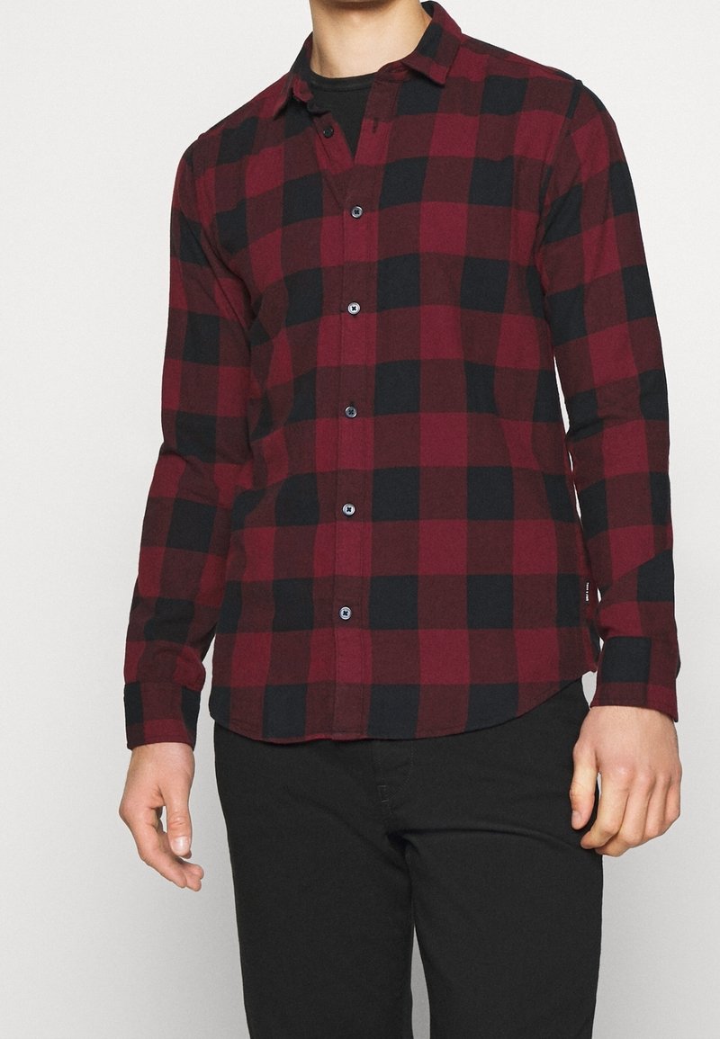 Chemise à carreaux rouge et noir avec un tissu doux, un col standard, des manches longues et des poignets à boutons. Boutons bleus et ourlet arrondi.