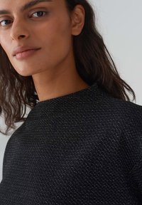 Schwarzes Top mit hohem Kragen, strukturierter Stoff mit metallischen Akzenten. Das Model zeigt ein natürliches Makeup und schulterlanges welliges Haar.