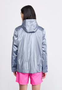 Silver waterproof jacket with a hood, featuring a textured finish and a lightweight design. Worn with bright pink shorts.