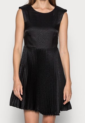 Femme portant une robe noire sans manches avec un motif texturé subtil et une jupe plissée, debout les bras détendus le long du corps.