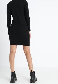 Robe noire en tricot côtelé avec manches longues, longueur au-dessus du genou, associée à des bottines noires à bout tirant et semelle épaisse.