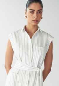 Weißes Hemdkleid mit vertikalen marineblauen Streifen, kurzen Ärmeln, einem Taillengürtel und einer Brusttasche. Glattes Material mit einer strukturierten Passform.