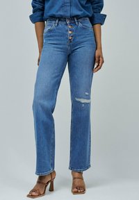 Jeans de mezclilla azul de tiro alto con pierna recta, que presentan un cierre de cinco botones, detalle rasgado en la rodilla y un ligero efecto deslavado.
