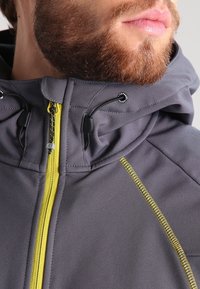 Homme barbu portant une veste à capuche grise avec une fermeture éclair jaune et un détail cousu jaune près de l'épaule.