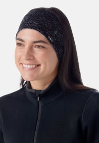 Femme souriante avec de longs cheveux bruns portant un bandeau noir à motif et un haut zippé noir, tournée légèrement vers la gauche sur un fond blanc.
