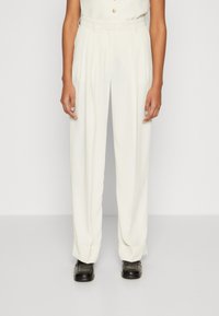 Pantalons blancs taille haute avec des plis, présentant une coupe ajustée et des ourlets retroussés. Le tissu a une texture lisse, complété par des chaussures noires.