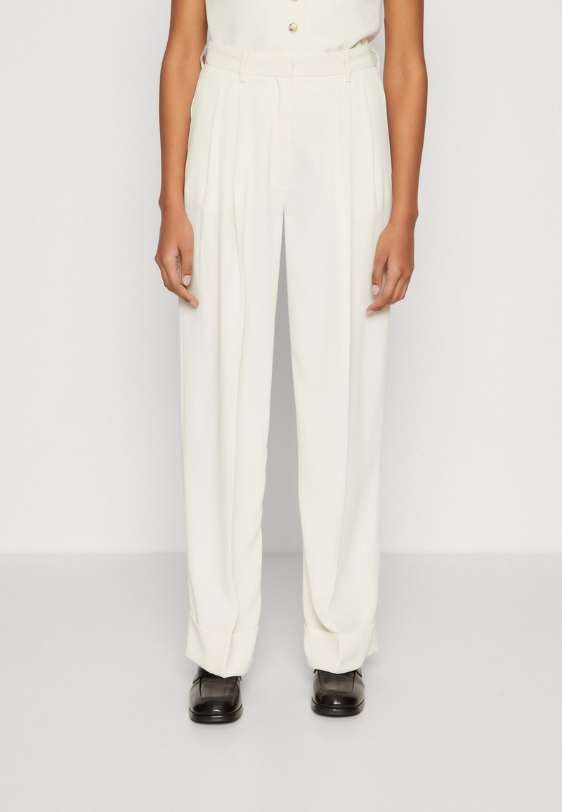 Pantalons blancs taille haute avec des plis, présentant une coupe ajustée et des ourlets retroussés. Le tissu a une texture lisse, complété par des chaussures noires.