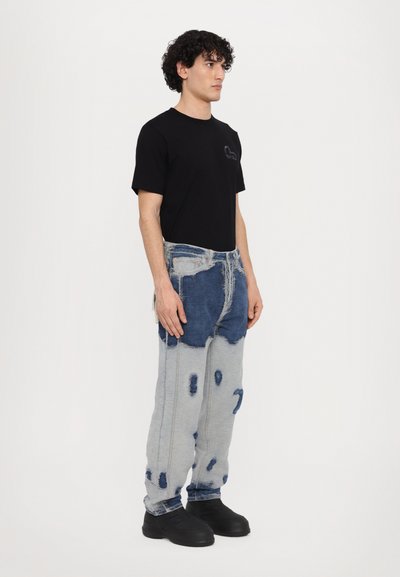 T-shirt en coton noir avec logo sur la poitrine, associé à un jean en denim usé présentant des zones de délavage clair et foncé ainsi qu'une forte décoloration, porté avec des chaussures noires.
