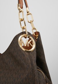 Sac à main en cuir marron avec des anses en chaîne dorée et un pendentif circulaire doré avec le logo MK suspendu près de la couture supérieure.