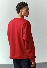 Rød sweatshirt med rund halsudskæring, dropped skuldre og ribbede manchetter. Stofmaterialet virker blødt med en glat overflade.