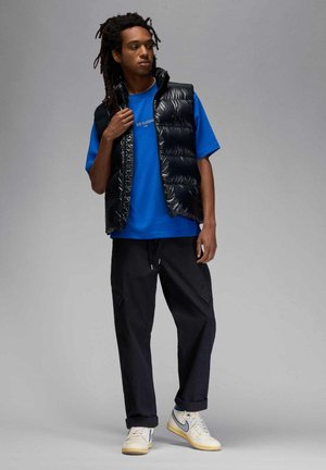 Joven con rastas que lleva un chaleco acolchado negro brillante sobre una camiseta azul de Air Jordan, pantalones negros y zapatillas blancas Nike.