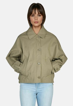 Young woman with short brown hair wearing light brown buttoned jacket and light blue jeans, hands in jacket pockets, neutral background.