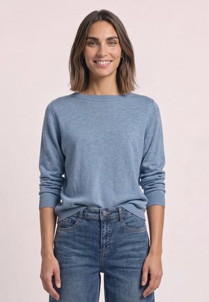 Femme aux cheveux bruns mi-longs portant un pull bleu clair et un jean bleu, souriante sur un fond clair uni.