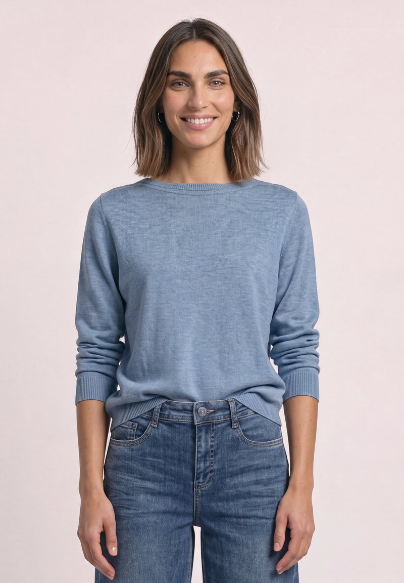 Femme aux cheveux bruns mi-longs portant un pull bleu clair et un jean bleu, souriante sur un fond clair uni.