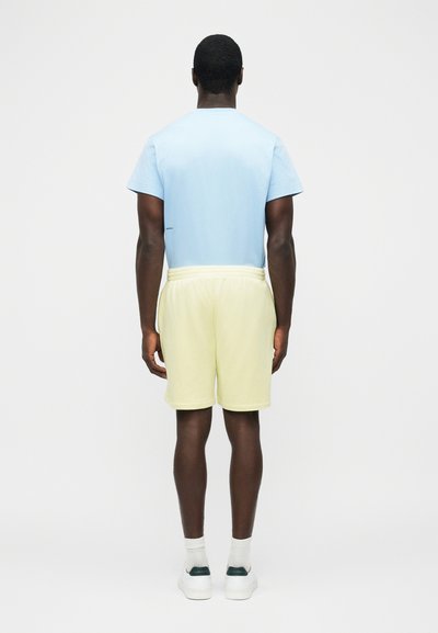 Man facing away wearing a light blue short-sleeve shirt, pale yellow shorts, white socks, and white sneakers on a plain white background.