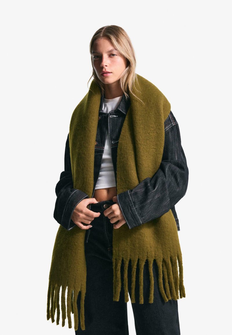 Limegrøn fransede tørklæde lavet af blødt uld. Modellen har det på over en sort denimjakke og en hvid top, hvilket fremhæver det teksturerede design.