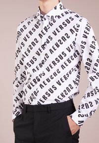 Witte overhemd met knopen met een gedurfde zwarte "VERSUS" print in verschillende hoeken, met een gladde textuur en zwarte knopen. Gecombineerd met zwarte broek.