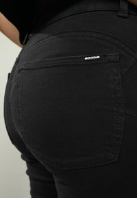 Jeans de mezclilla negra con una vista en primer plano que resalta el bolsillo trasero y el detalle del logo metálico. La tela parece suave con costuras sutiles.