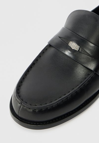 Mustad mustad nahast loafers, millel on sile viimistlus, õmmeldud detailid ja horisontaalne riba koos dekoreeritud metall-embleemiga.