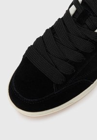 Acercamiento a una zapatilla de gamuza negra con cordones gruesos negros y suela de goma blanca sobre un fondo liso.