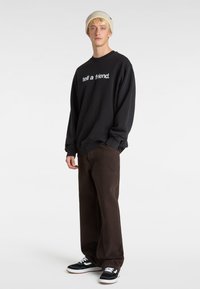 Sort sweatshirt med teksten "fortæl en ven." kombineret med mørkebrune bukser og sorte sneakers med hvide detaljer. Modellen bærer en beige beanie.