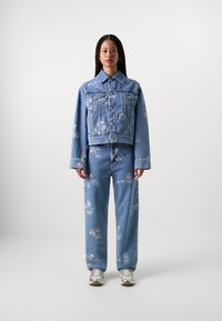Ensemble en denim léger bleu, comprenant une veste et un pantalon, avec des motifs imprimés blancs. La veste a un design court avec des fermetures à boutons.