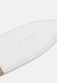 Étiquette en cuir blanc avec le texte "DKNY" en relief, bordée de tissu beige. Présente des coutures précises le long du bord, texture lisse.
