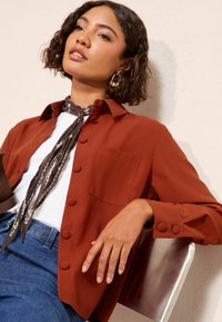 Rozsda színű, galléros ing, gomb részletekkel és elülső zsebbel, fehér felsővel és sötét farmernadrággal párosítva. Puha szövet textúra.