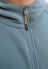 Hoodie zippé bleu clair avec une capuche côtelée texturée, tirette de fermeture éclair orange contrastante et tissu doux, présentant une coupe standard et un design sans coutures.