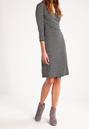 Femme portant une robe noire et blanche à motif, longueur genou, manches trois-quarts, et des bottines en daim gris sur un fond uni.