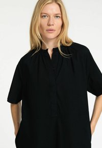 Černá košile s krátkým rukávem, s zapínáním na knoflíky u krku, s nařasenými detaily a volným střihem. Vyrobena z hladkého materiálu s bočními kapsami.