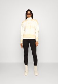 Sweatshirt tie-dye branco e amarelo com um pescoço alto, punhos e barra canelados, combinado com leggings pretas e botas de plataforma em tom creme.