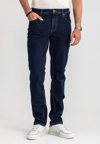 Ciemnoniebieskie dżinsy z denimu o wąskim kroju, z pomarańczowymi szwami, zapięciem na guzik i zamek błyskawiczny oraz przednimi kieszeniami. Połączone z białymi trampkami.