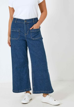 Weite blaue Jeans mit hohem Bund, zwei großen vorderen Taschen und Kontrastnähten, kombiniert mit weißen Turnschuhen.
