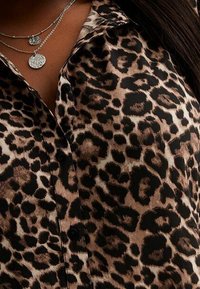 Blusa con stampa leopardata che presenta una combinazione cromatica marrone e nera, con una chiusura a bottoni sul davanti e una texture liscia. Collana a strati in argento visibile.