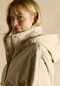 Jachetă impermeabilă bej cu guler înalt și glugă ajustabilă. Include un fermoar frontal și detalii de cusătură pe cusătura umărului.