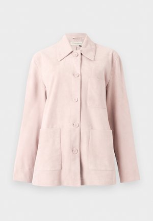 Femeie cu păr lung și închis la culoare poartă o jachetă roz pal cu nasturi, blugi închiși la culoare, balerini negri și ține o geantă neagră, pe fundal alb.