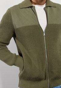 Olivgrön zip-sweater med stickad textur på kroppen och slät tyg på toppen. Har en krage och sidofickor.
