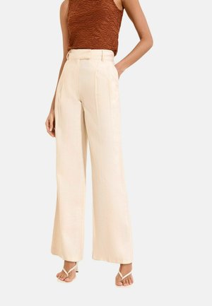 Woman wearing high-waisted wide-leg cream trousers, textured sleeveless brown top, and white heeled sandals, standing with one hand in pocket.