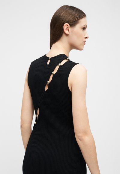 Femme aux cheveux bruns raides portant une robe noire sans manches en maille côtelée avec des accents de boutons dorés diagonaux dans le dos, sur un fond uni.