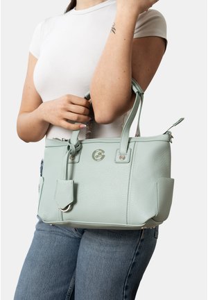 Frau in weißem T-Shirt und blauer Jeans hält eine hellblaue Lederhandtasche mit doppelten Griffen und vorderem Logoverschluss.