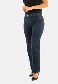 Pantalones vaqueros de mezclilla azul oscuro con corte recto, que presentan bolsillos frontales y cierre de botón, combinados con tacones altos negros.