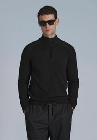 Schwarzer Zip-Kragen-Pullover mit slim fit, aus weichem Stoff, verfügt über Raglanärmel und einen gerippten Saum. Getragen mit schwarzen Jogginghosen mit Kordelzug.