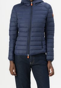 Person in einer marineblauen gesteppte Jacke mit Reißverschluss und Kapuze sowie dunklen Jeans, der mit entspannten Armen an den Seiten vor einem weißen Hintergrund steht.