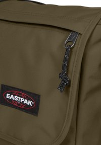 Olivově zelená taška Eastpak s texturovanou tkaninou, zapínáním na zip a černou nášivkou s červenými akcenty. Přítomen černý šňůrkový detail.