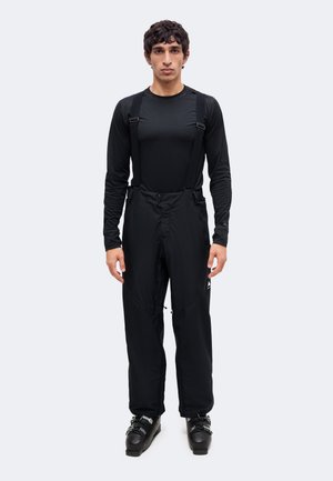 Hombre con pantalones de esquí negros con tirantes, camiseta negra de manga larga y botas de esquí negras, de pie frente a un fondo claro y liso.