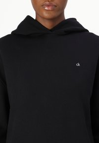 Sweatshirt negru cu glugă, confecționat dintr-un material moale, cu o croială relaxată, având un mic logo alb "ck" pe piept. Textură netedă, fără modele.
